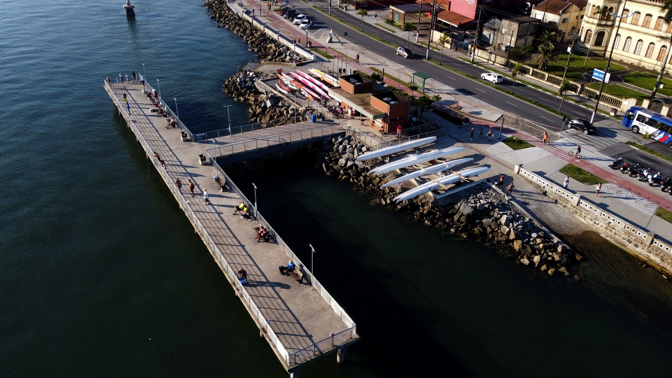 Vista aérea do Deck do Pescador, em Santos, mostrando o píer sobre o mar, pessoas caminhando e embarcações de remo alinhadas na área próxima