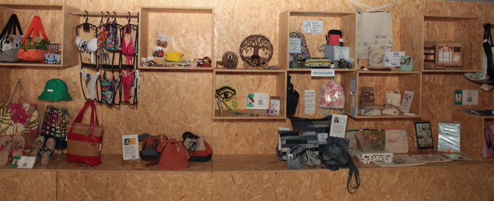 Parede expositiva com nichos de madeira exibindo bolsas, sapatos, chapéus e objetos decorativos, em ambiente organizado e voltado ao artesanato