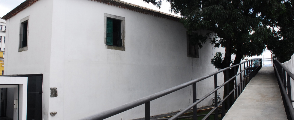 Prédio branco com janelas verdes ao lado de rampa de concreto com corrimão metálico, sombreado por uma grande árvore em cenário urbano