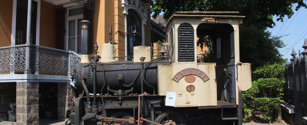 Uma foto mostra uma pequena locomotiva a vapor antiga, preta e bege claro, com a inscrição "ITANGUÁ" e o número 1, estacionada ao ar livre em frente a um prédio de cor ocre e varanda de ferro