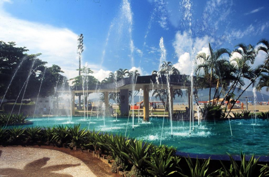 A imagem mostra a Fonte do Sapo, um dos pontos mais tradicionais da orla de Santos. No centro, jatos de água se elevam de um grande espelho d’água cercado por plantas ornamentais e palmeiras, criando um cenário refrescante e agradável. Ao fundo, é possível ver a praia, com barracas e guarda-sóis coloridos, além do mar e do céu azul pontilhado por nuvens claras. A estrutura da fonte, com colunas e áreas sombreadas, convida moradores e turistas a fazerem uma pausa durante o passeio pela orla.
