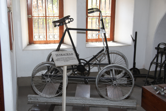 Tetraciclo antigo exposto em um museu, feito de metal com estrutura simples e quatro rodas finas semelhantes às de bicicleta. Está posicionado sobre uma base de madeira, com uma placa explicativa à frente