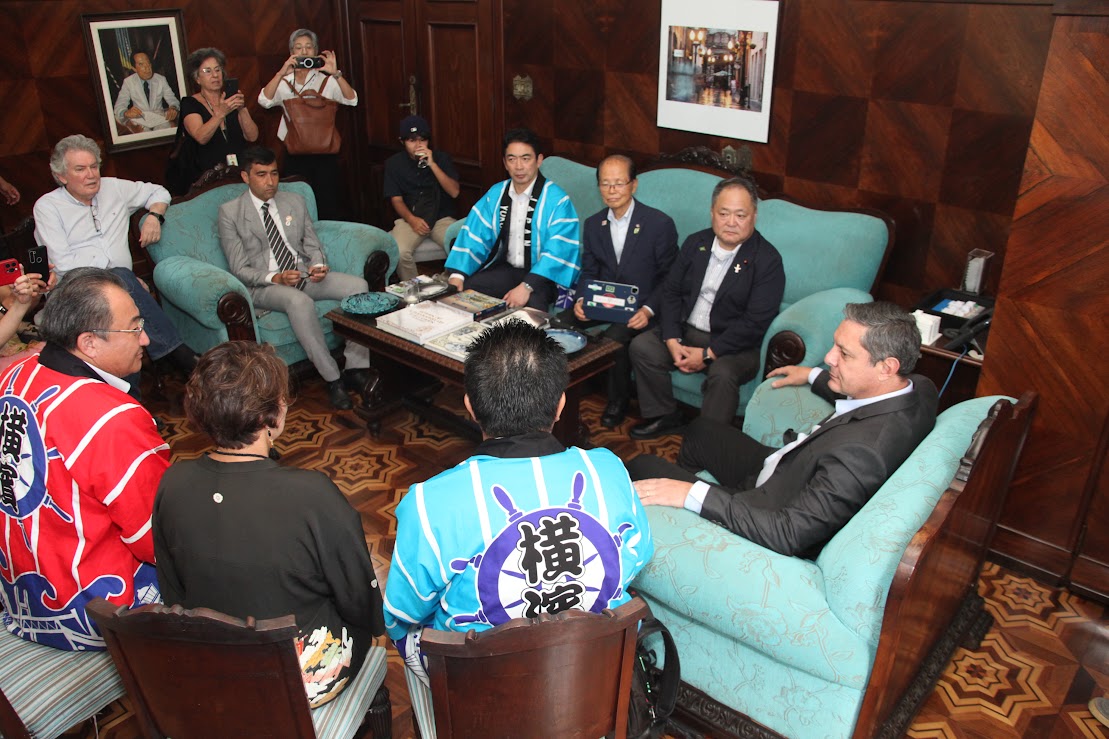 Reunião formal e intercultural ocorre em uma sala com painéis de madeira e móveis azuis onde vários indivíduos, alguns vestindo casacos japoneses tradicionais happi vermelhos e azuis e outros de terno, estão sentados em torno de uma mesa de centro