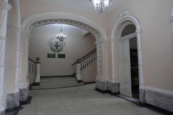 Hall de entrada clássico com piso de mármore e paredes bege, destacando uma escadaria lateral com corrimão branco e pilares ornamentados. Um brasão pintado e lustres suspensos marcam a transição para a escada principal.