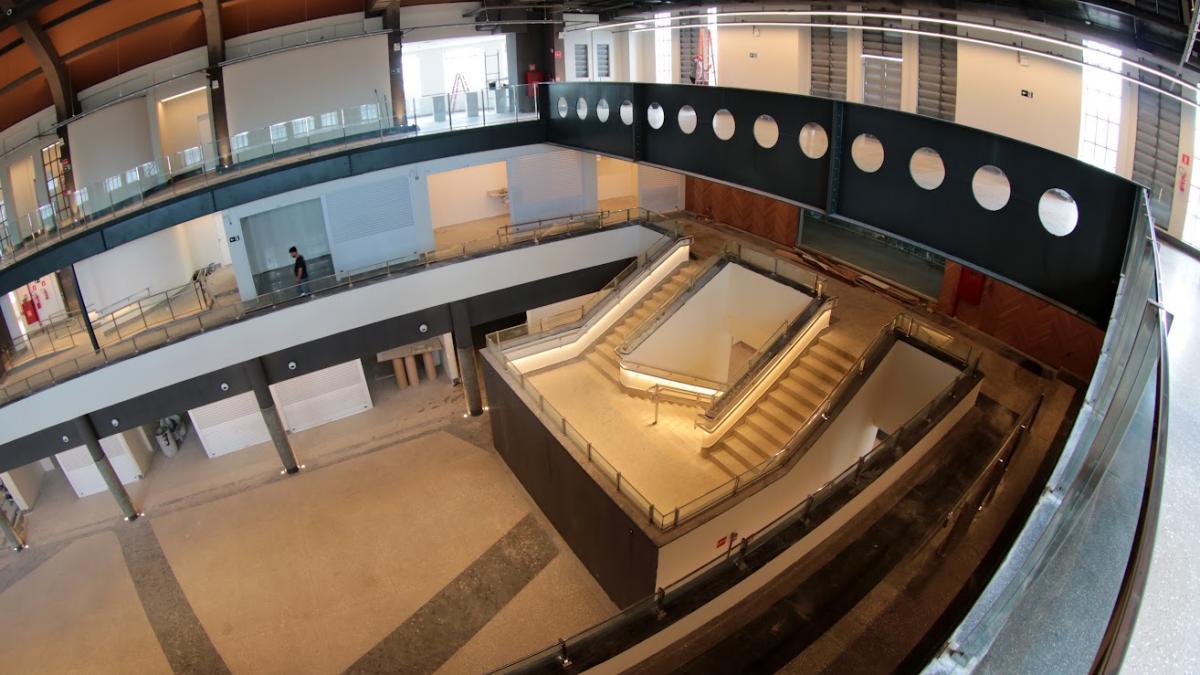 Imagem de ângulo superior de um interior moderno e espaçoso em construção ou renovação, apresentando um grande átrio central com escadas de acesso e uma passarela metálica superior com aberturas circulares
