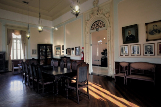Sala de reunião com uma longa mesa de madeira escura e cadeiras almofadadas em primeiro plano, sobre um piso de madeira brilhante. A sala é decorada com retratos nas paredes, uma estante envidraçada e lustres antigos, com luz solar entrando por uma janela gótica.