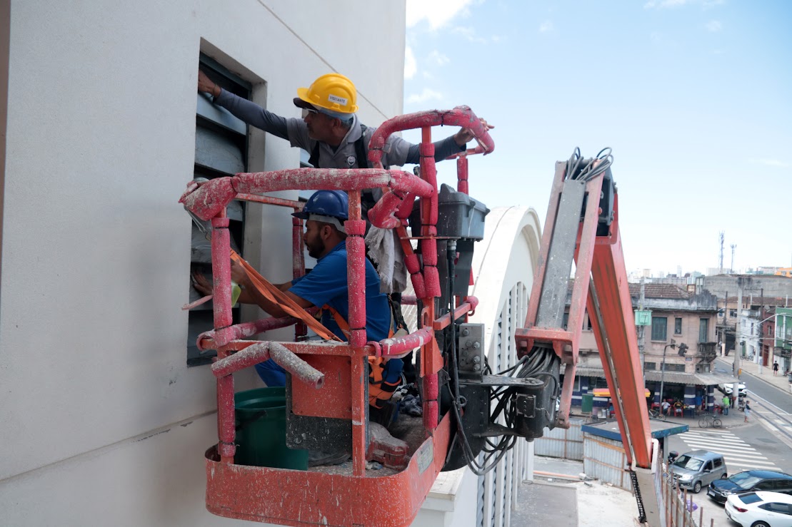 Dois trabalhadores da construção civil, usando capacetes de segurança, estão em uma plataforma elevatória vermelha realizando trabalhos de manutenção ou instalação na fachada de um edifício, com a paisagem urbana ao fundo