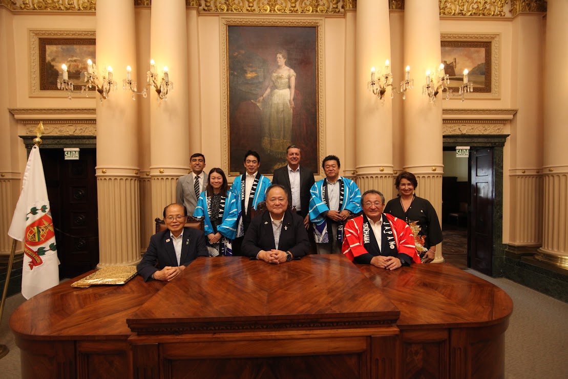 Um grupo de oito pessoas, algumas vestindo casacos japoneses tradicionais happi azuis e vermelhos e outras de trajes de negócios, estão posando para uma foto atrás de uma grande mesa de madeira em uma sala formal com colunas e um quadro emoldurado na parede