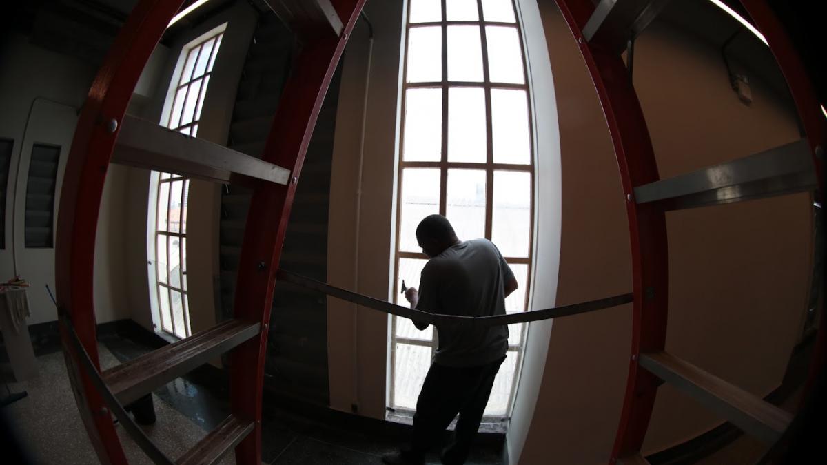 Uma imagem de ângulo distorcido mostra um homem de costas, realizando reparos em uma grande janela vertical com caixilhos de madeira