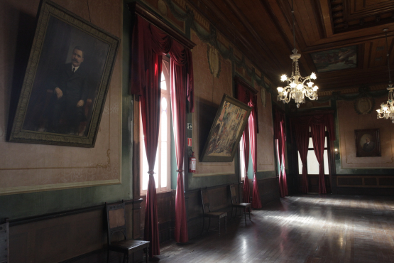 Grande salão de festas com piso de madeira, teto alto e janelas longas adornadas com cortinas de veludo vermelho. As paredes em dois tons exibem grandes retratos inclinados e lustres de cristal pendurados.