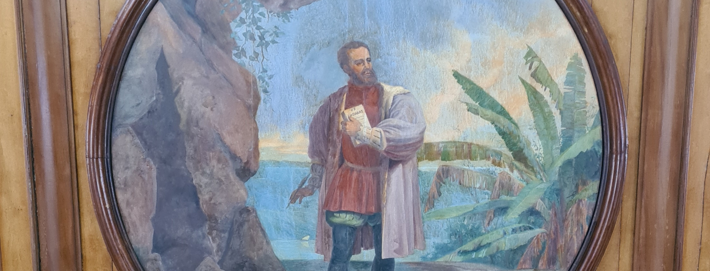 Pintura oval em moldura de madeira, retratando um homem barbudo em vestes históricas (camisa vermelha e casaco roxo) em um cenário costeiro. Ele segura um pergaminho com a palavra "Lusíadas", sugerindo ser Luís Vaz de Camões, em frente a uma gruta e folhagens tropicais.