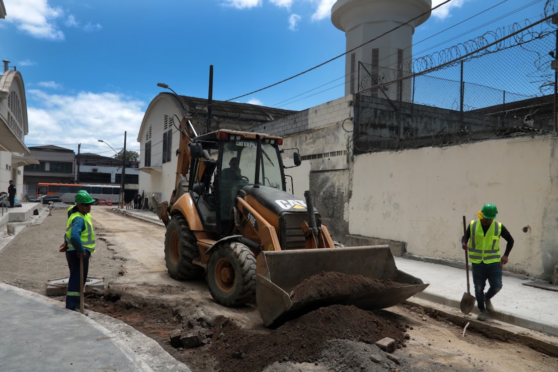 Uma retroescavadeira amarela e preta está em uma rua em construção ou reparo, com terra e cascalho no solo, enquanto dois trabalhadores de colete e capacete verde supervisionam ao redor da máquina