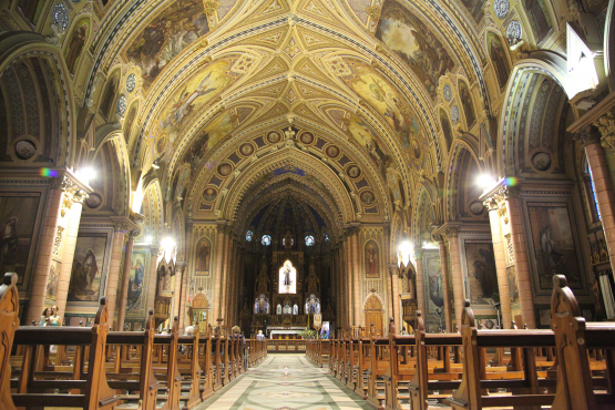 Interior da Basílica do Embaré em estilo neogótico, com uma longa nave central ladeada por bancos de madeira e colunas. O teto em abóbada é ricamente decorado com afrescos e ornamentos dourados, iluminado por luzes internas que realçam os detalhes