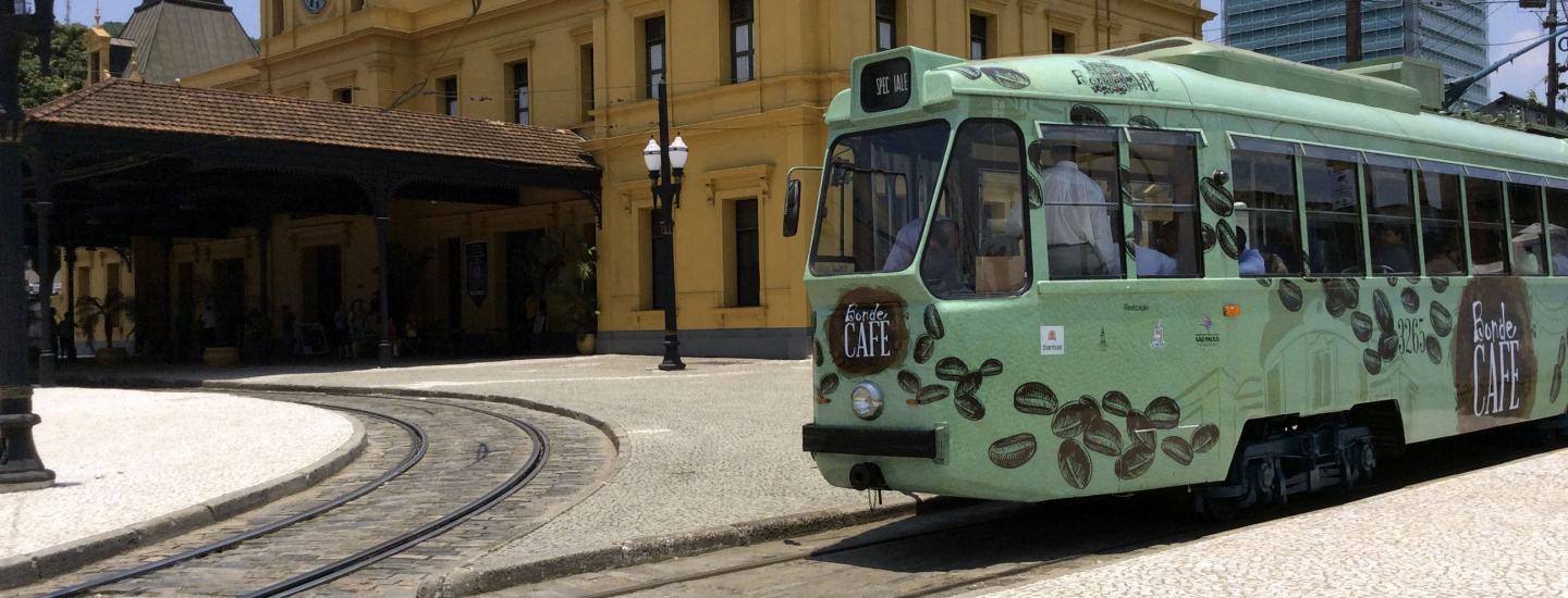Bonde verde decorado com grãos de café passa em frente a um prédio histórico amarelo, sob céu ensolarado. Pessoas estão dentro do bonde e próximas à estação.