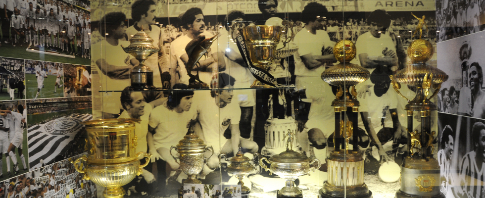 Uma coleção de troféus dourados em uma vitrine, com diversas taças de futebol. Ao fundo, uma grande imagem em sépia mostra um time de futebol, possivelmente o Corinthians, comemorando
