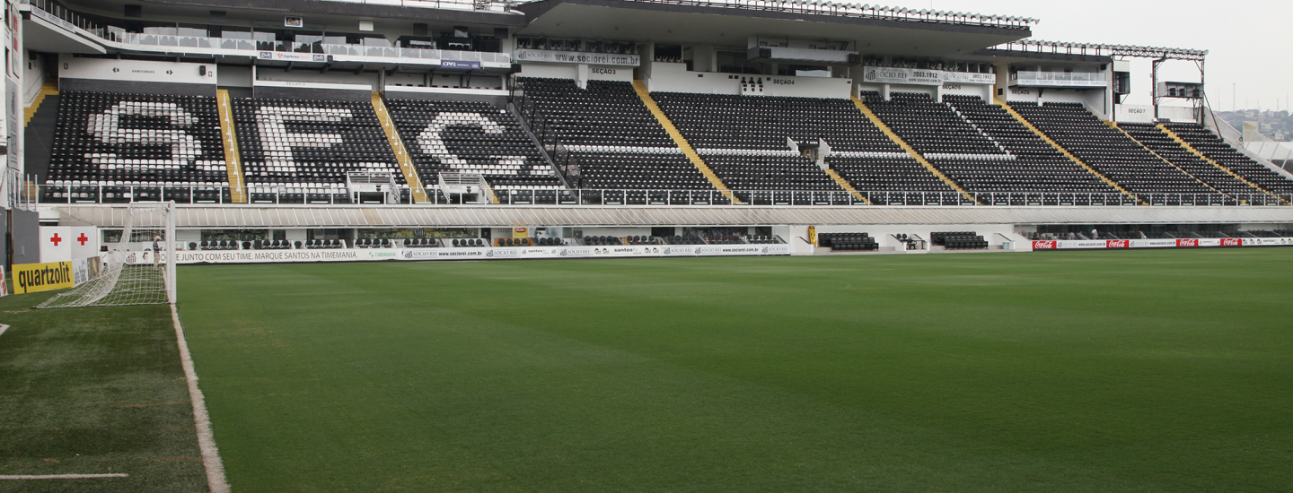 Campo de futebol com gramado verde impecável, visto de perto, com a arquibancada ao fundo exibindo as letras “SFC” em assentos pretos e brancos. A cena mostra parte do gol e a estrutura do estádio vazia