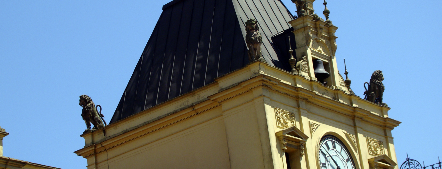 Fachada do edifício em tom amarelo claro com telhado escuro, um relógio na torre e estátuas de leões nos cantos contra um céu azul brilhante
