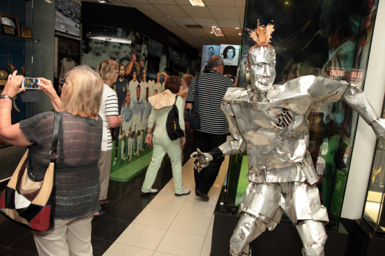Visitantes caminham por um corredor de museu esportivo enquanto observam fotos e painéis; à frente, uma escultura metálica em forma de jogador de futebol chama a atenção. Uma mulher fotografa a cena, destacando a interação do público com o espaço.