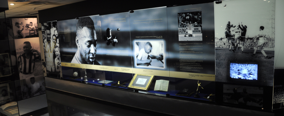 Painel expositivo com fotos em preto e branco de um jogador de futebol lendário, acompanhado de textos, objetos históricos e pequenos troféus em vitrines iluminadas. A mostra apresenta imagens de partidas, momentos íntimos e registros de sua trajetória esportiva.