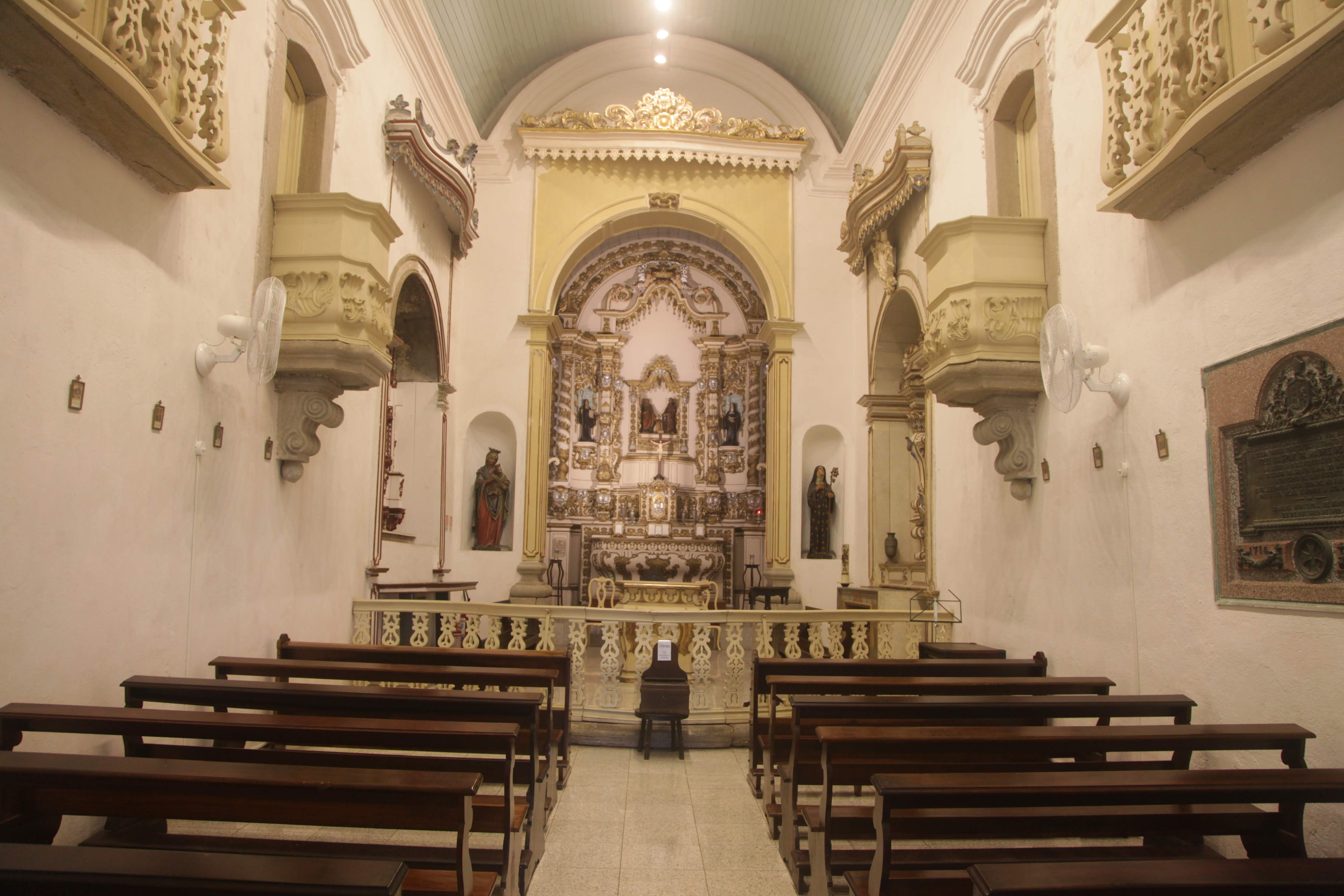 Uma fotografia interna simétrica mostra a nave central de uma capela colonial simples, com longas fileiras de bancos de madeira escura e um altar ricamente trabalhado em talha dourada e branca ao fundo