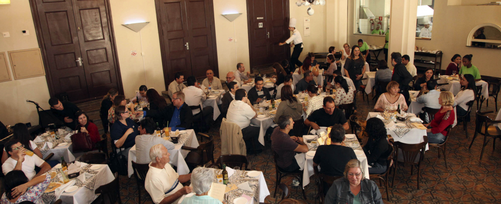 Restaurante movimentado com várias mesas ocupadas por pessoas almoçando e conversando em ambiente acolhedor e iluminado. Garçons circulam entre as mesas atendendo os clientes