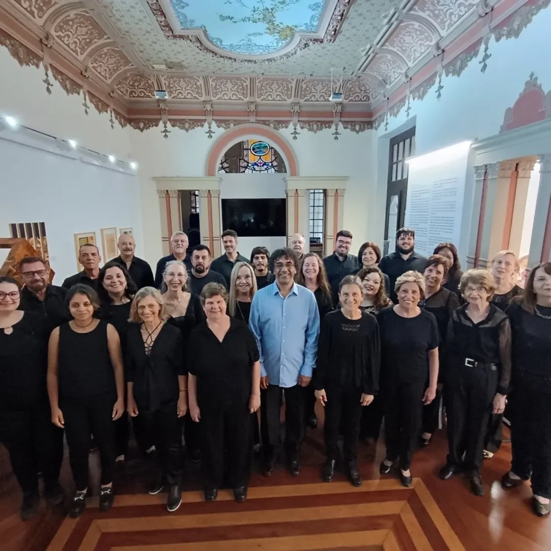 Foto de um grupo de pessoas de um coral, vestindo preto, sorrindo e posando em um salão ornamentado
