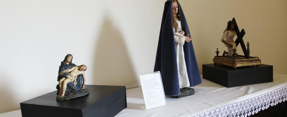 Uma fotografia exibe um conjunto de três estátuas de arte sacra expostas sobre uma mesa coberta com uma toalha branca, destacando-se uma escultura maior da Virgem Maria envolta em um manto azul, ao lado de uma Pietà e uma representação de Cristo carregando a cruz