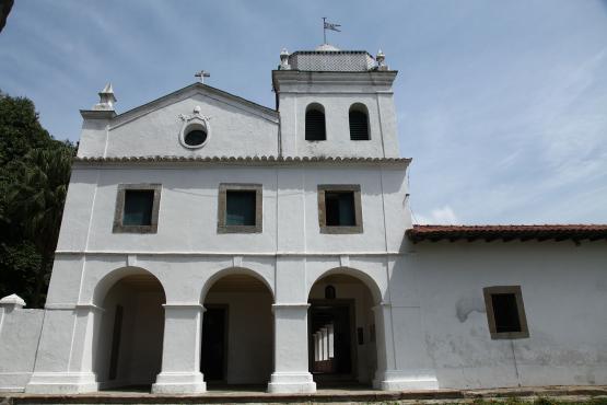 Uma fotografia externa de uma igreja ou capela colonial branca de arquitetura simples, apresentando uma fachada com três arcos na parte inferior e uma torre sineira no lado direito, sob um céu parcialmente nublado