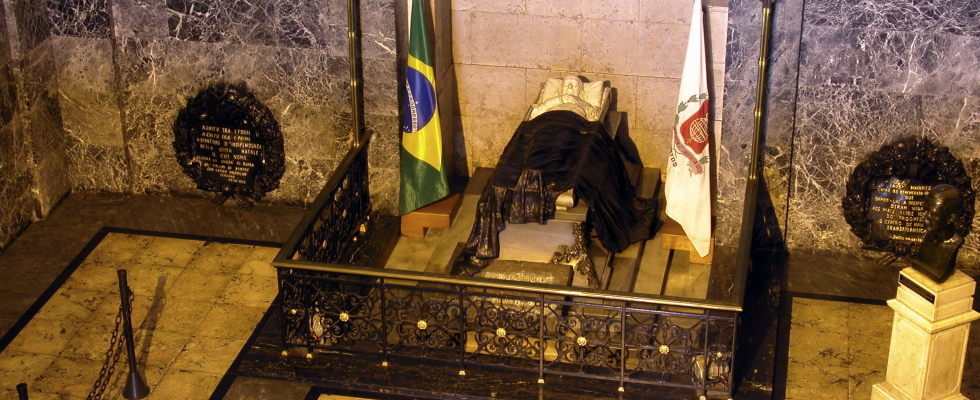 A top-down view of a monumental tomb or sarcophagus covered with black fabric and surrounded by a wrought iron railing, flanked by the flags of Brazil and São Paulo, in a setting lined with dark marble and commemorative plaques
