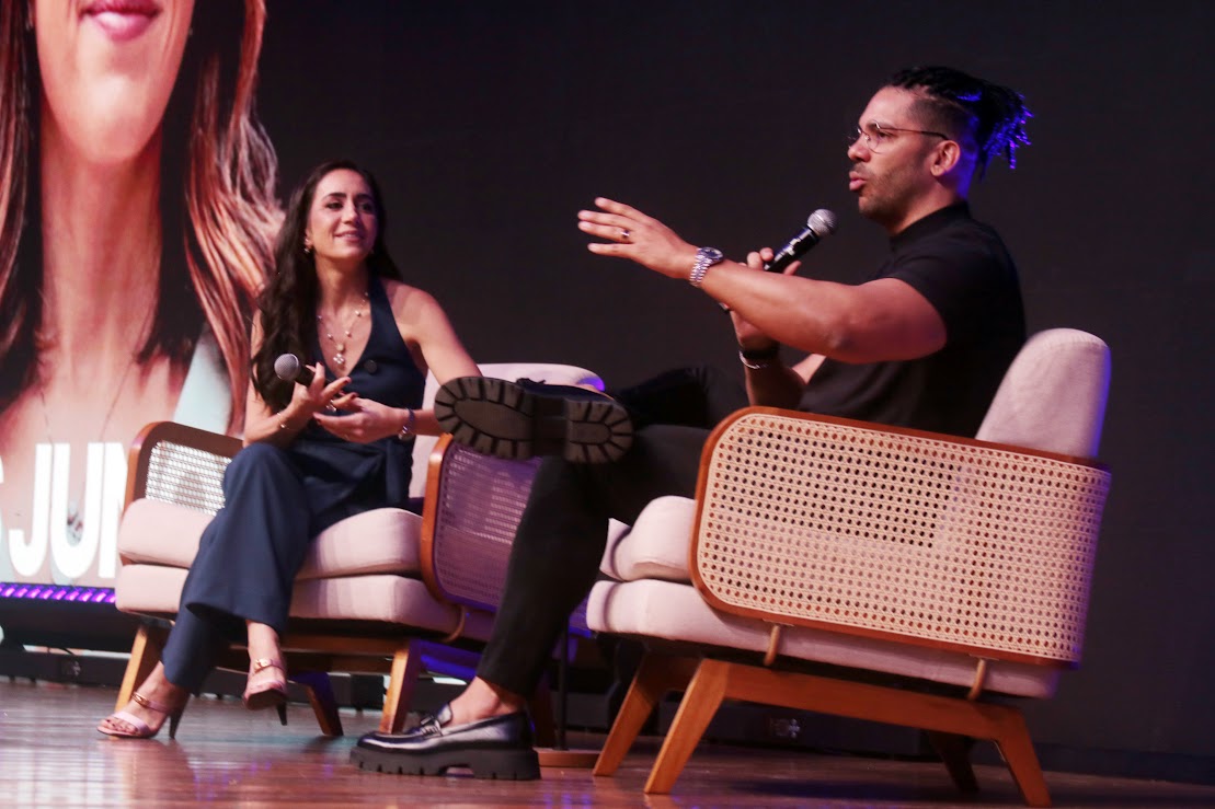 Um homem e uma mulher sentados em poltronas de vime em um palco estão participando de um painel ou entrevista, ambos segurando microfones enquanto o homem gesticula