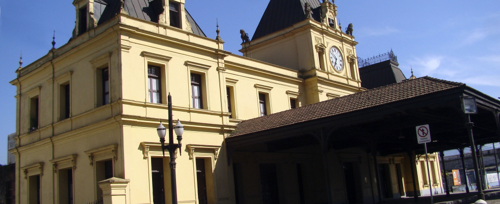 Prédio histórico amarelo com torre central e relógio na fachada, telhado escuro e detalhes arquitetônicos clássicos. O céu está azul e claro, destacando a construção