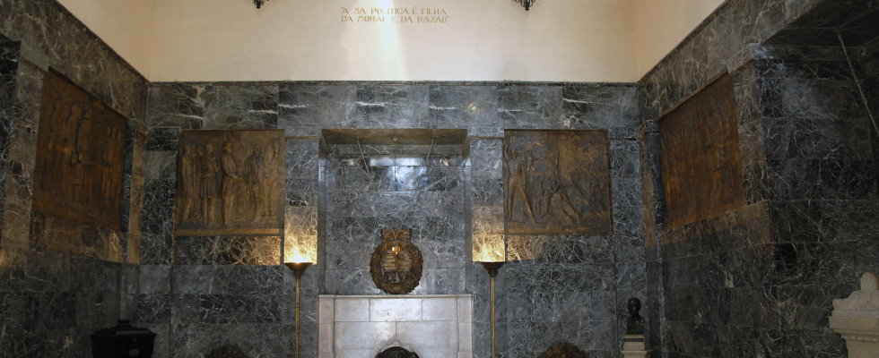 Uma foto de uma sala solene com paredes revestidas de mármore escuro, apresentando quatro painéis em relevo/bronze emoldurados, uma lareira central de mármore claro com um emblema redondo acima, e uma inscrição no alto da parede, iluminada por tochas laterais