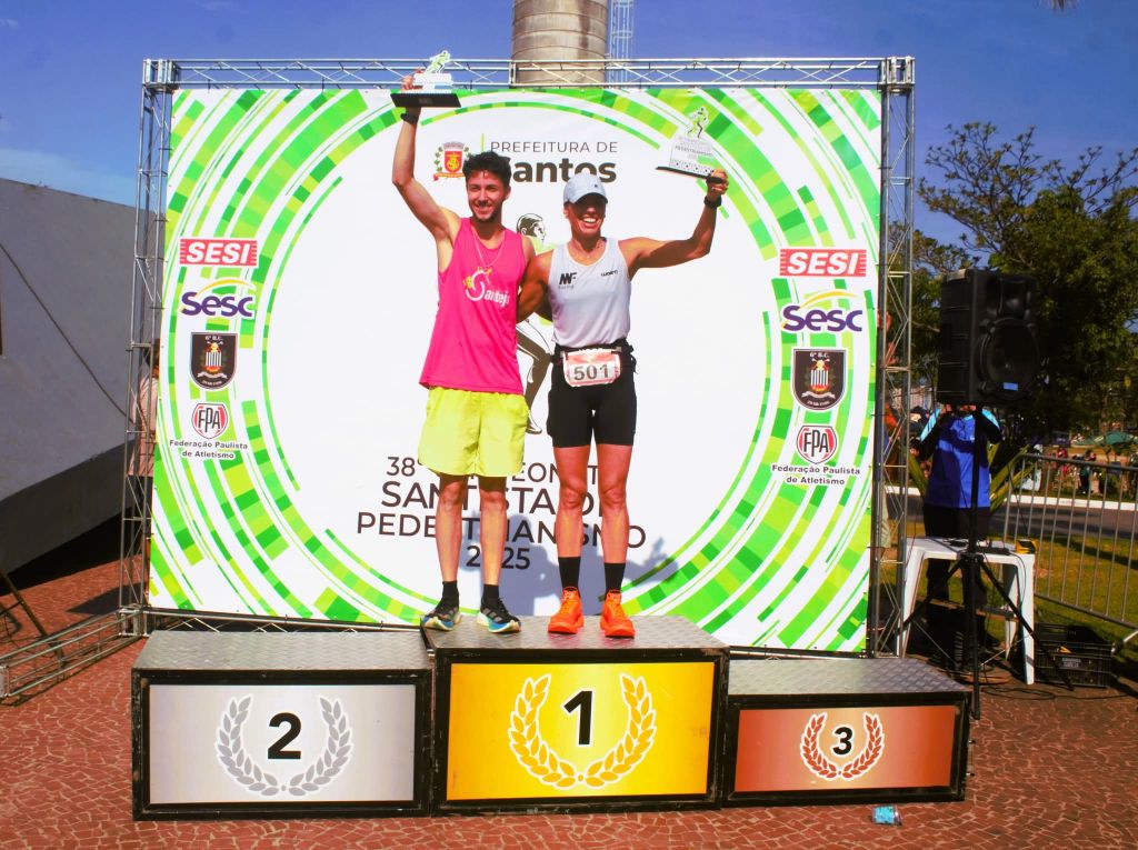 Dois atletas vencedores, um homem e uma mulher, celebram no pódio de uma corrida com troféus em mãos, em frente a um banner com o logotipo da Prefeitura de Santos