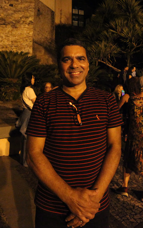 Um homem de meia-idade, vestindo uma camisa listrada em vermelho e preto, sorri para a câmera em uma área externa à noite, com plantas e outras pessoas desfocadas ao fundo