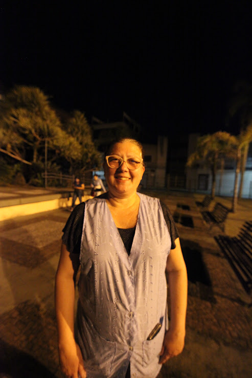 Uma mulher sorridente com óculos e um colete claro posa para a foto à noite em uma praça, com iluminação focal e fundo escuro