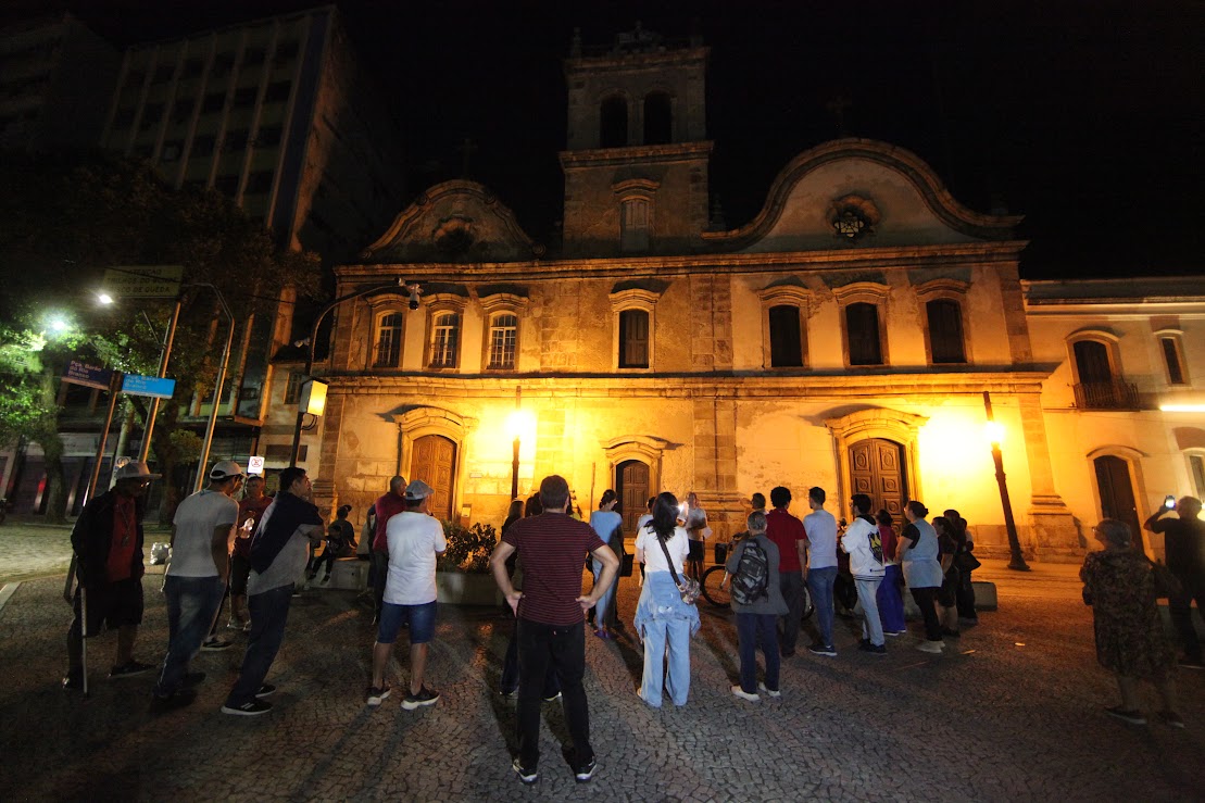 Um grupo de pessoas está reunido à noite em frente a uma imponente e iluminada igreja histórica