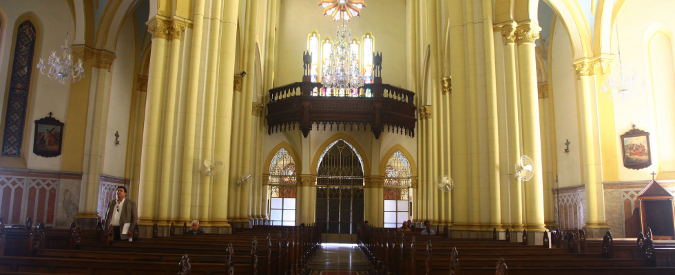 The image depicts the interior of a large church with distinctly Neo-Gothic architecture, evidenced by the verticality of its elements and pointed arches. The eye is guided along the central axis of the nave, flanked by long rows of dark wood pews, which reinforce the symmetry and the sense of depth of the space.