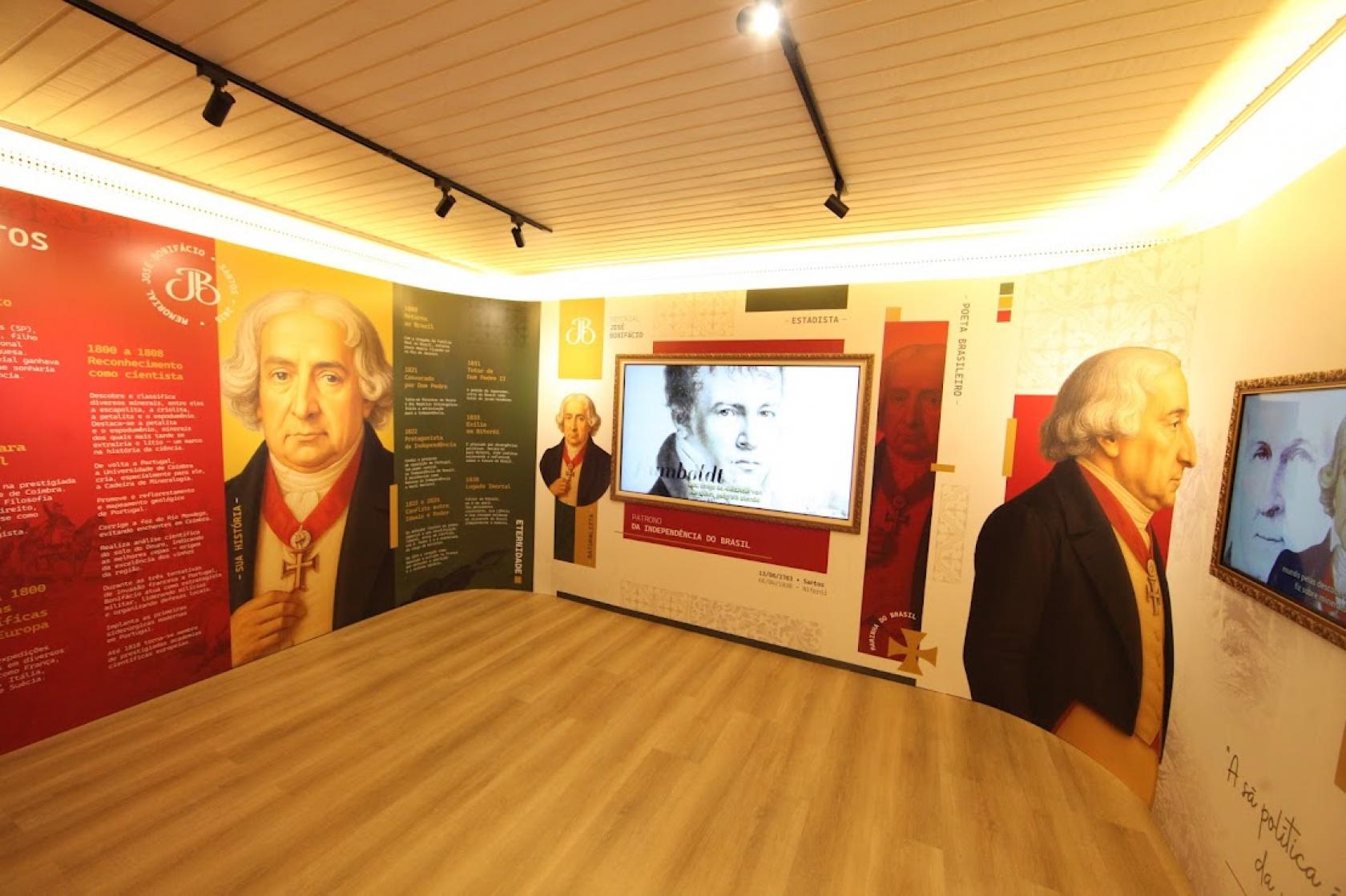 A imagem mostra uma sala de exposição moderna, bem iluminada, dedicada à vida e trajetória de José Bonifácio de Andrada e Silva. As paredes são totalmente envelopadas por painéis coloridos — em tons de vermelho, verde, amarelo e branco — que apresentam textos informativos, datas importantes e títulos que contam diferentes fases de sua atuação, como estadista, cientista e patriota da Independência do Brasil.