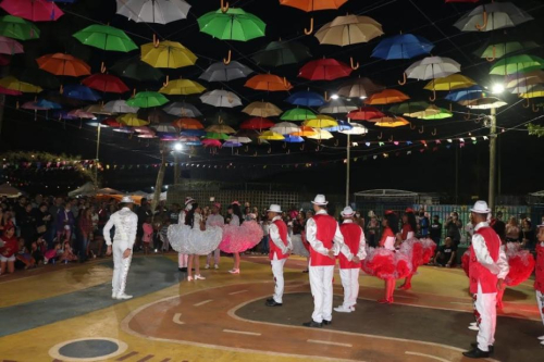 Um grupo de dançarinos de quadrilha se apresenta à noite para o público sob uma decoração repleta de guarda-chuvas coloridos