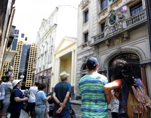 Na imagem, um grupo de pessoas participa de um walking tour no Centro Histórico de Santos. Elas estão reunidas em uma rua de paralelepípedos, observando atentamente a fachada de um prédio histórico de estilo clássico, com detalhes ornamentados e um medalhão esculpido acima da entrada. Os participantes, vestidos de forma casual, alguns com chapéus e câmeras, demonstram interesse enquanto recebem informações sobre a história e a arquitetura local.