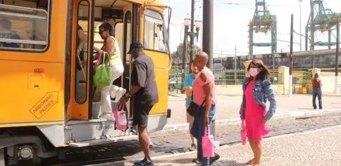 A imagem mostra um grupo de pessoas embarcando em um bonde amarelo, em um ambiente ensolarado. O veículo tem um visual clássico e está parado em um ponto de embarque pavimentado com pedras.  À frente, uma mulher de vestido rosa e jaqueta jeans, usando máscara facial, aguarda sua vez de subir. Outras pessoas já estão entrando ou próximas à porta, carregando bolsas coloridas — algumas em tons de rosa e verde.