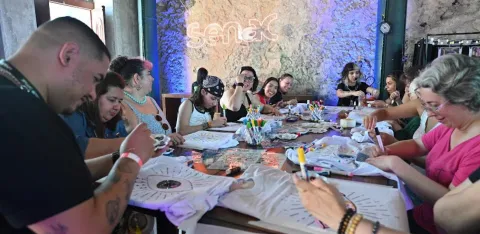 Um grupo numeroso de pessoas está reunido em torno de uma mesa retangular, participando de uma oficina criativa em um ambiente interno com paredes de pedra e iluminação decorativa em tons de roxo e azul. Ao fundo, destaca-se o letreiro iluminado com a palavra “Senac”, indicando a parceria ou local da atividade.  Os participantes — de diferentes idades — estão concentrados e sorridentes, personalizando camisetas com canetas coloridas e outros materiais de arte.