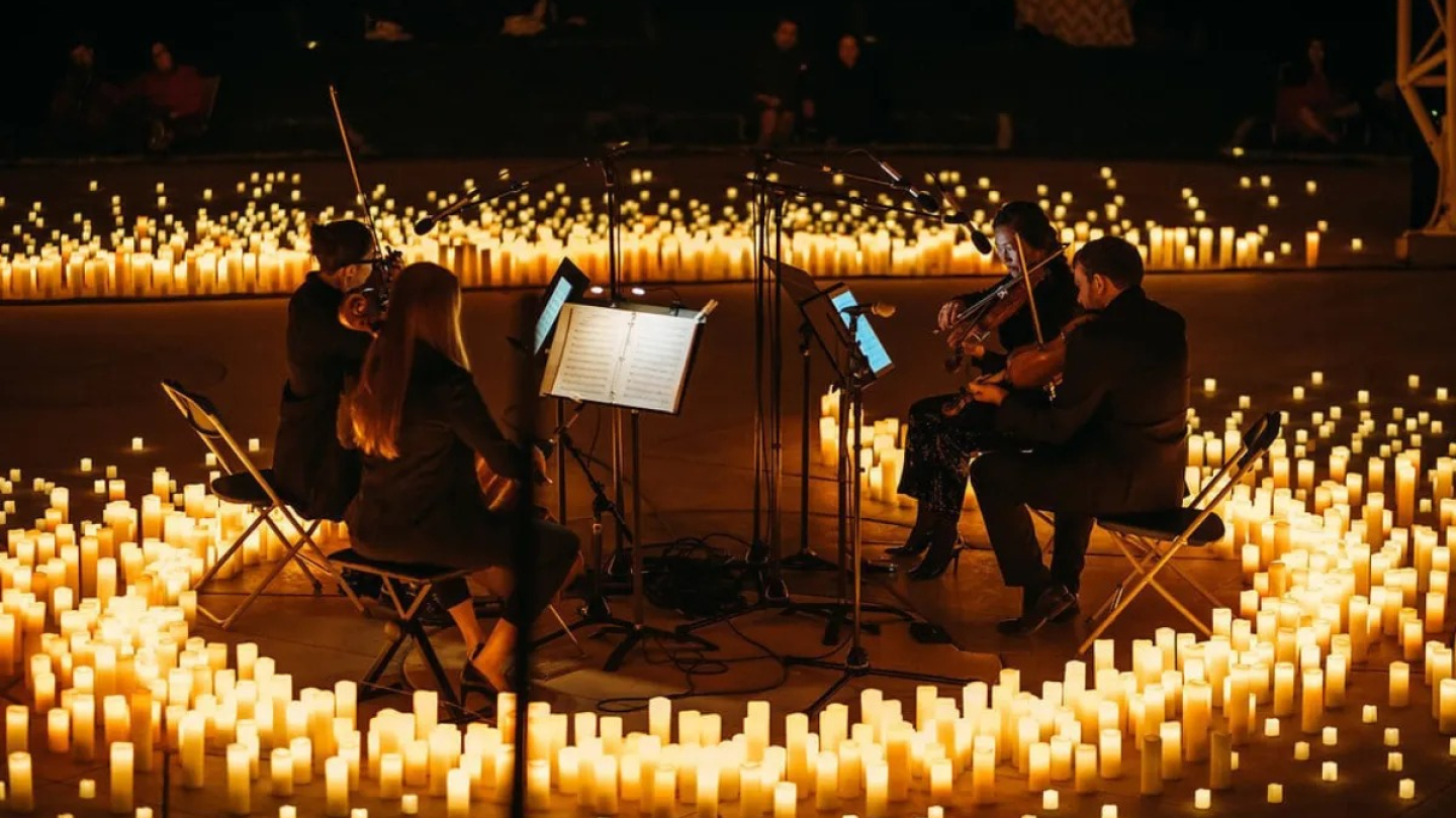Quatro músicos eruditos se apresentam em um palco à noite, cercados por centenas de velas acesas no chão e em plataformas, criando uma atmosfera aconchegante e intimista
