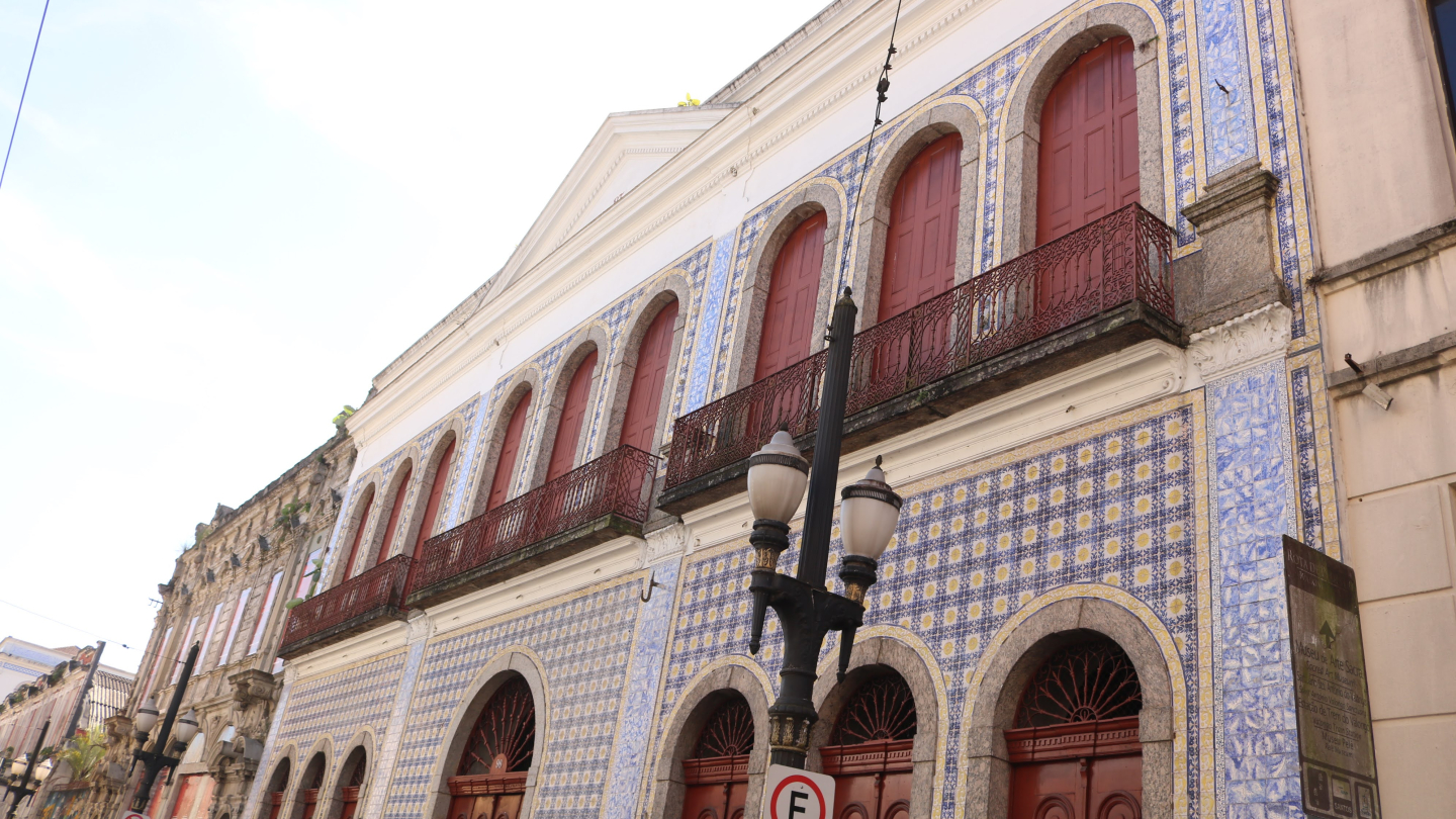 A fachada de um prédio histórico de dois andares, com a frente coberta por azulejos brancos e azuis. No andar de cima há janelas com venezianas e portas de madeira avermelhada, e no de baixo há arcos com portas de madeira. A frente é adornada com sacadas de ferro forjado e há um poste de luz na frente.