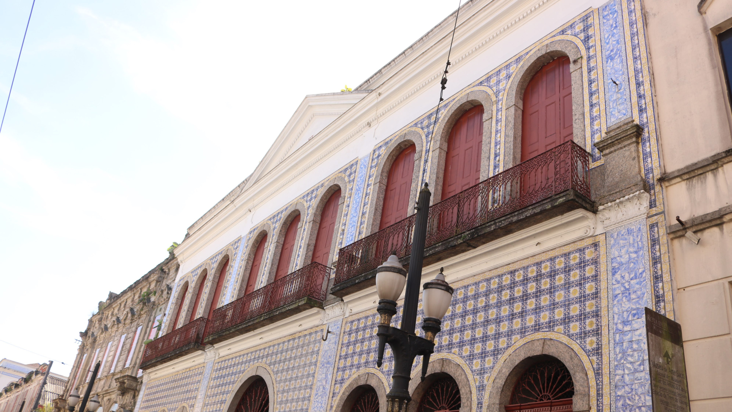 A fachada de um prédio histórico de dois andares, com a frente coberta por azulejos brancos e azuis. No andar de cima há janelas com venezianas e portas de madeira avermelhada, e no de baixo há arcos com portas de madeira. A frente é adornada com sacadas de ferro forjado e há um poste de luz na frente.