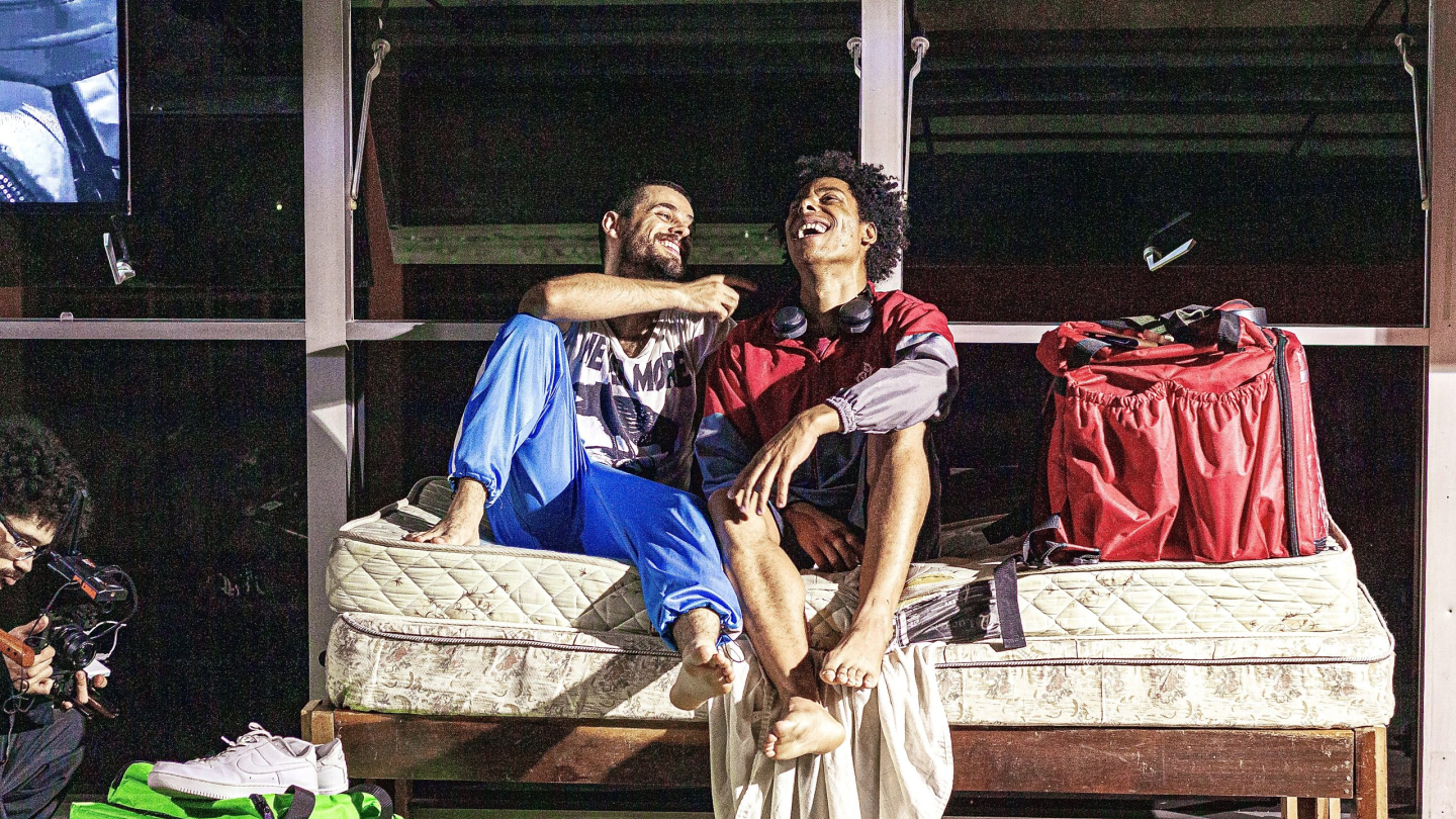 Dois homens sentados em um colchão, rindo juntos, em um cenário simples com mochilas de entregador ao lado.