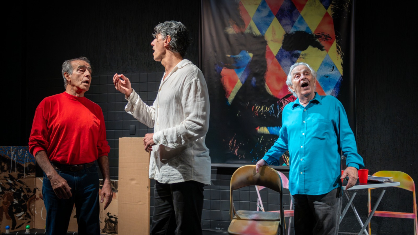 Três atores se apresentam no palco, em frente a um painel colorido com rosto estilizado de Plínio Marcos ao fundo