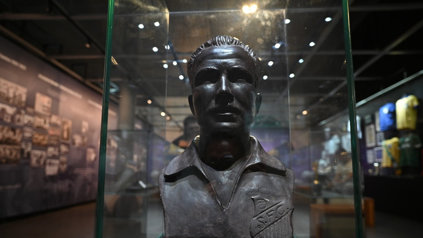 A imagem mostra um busto de bronze de José Macia, mais conhecido como Pepe, em exibição no Museu Pelé, como parte da exposição sobre sua trajetória no futebol. O busto está protegido por uma vitrine de vidro, destacando-se pela forma detalhada do rosto de Pepe, com uma expressão séria e imponente. Ele usa a camisa do Santos Futebol Clube, com o escudo do clube visível no busto.