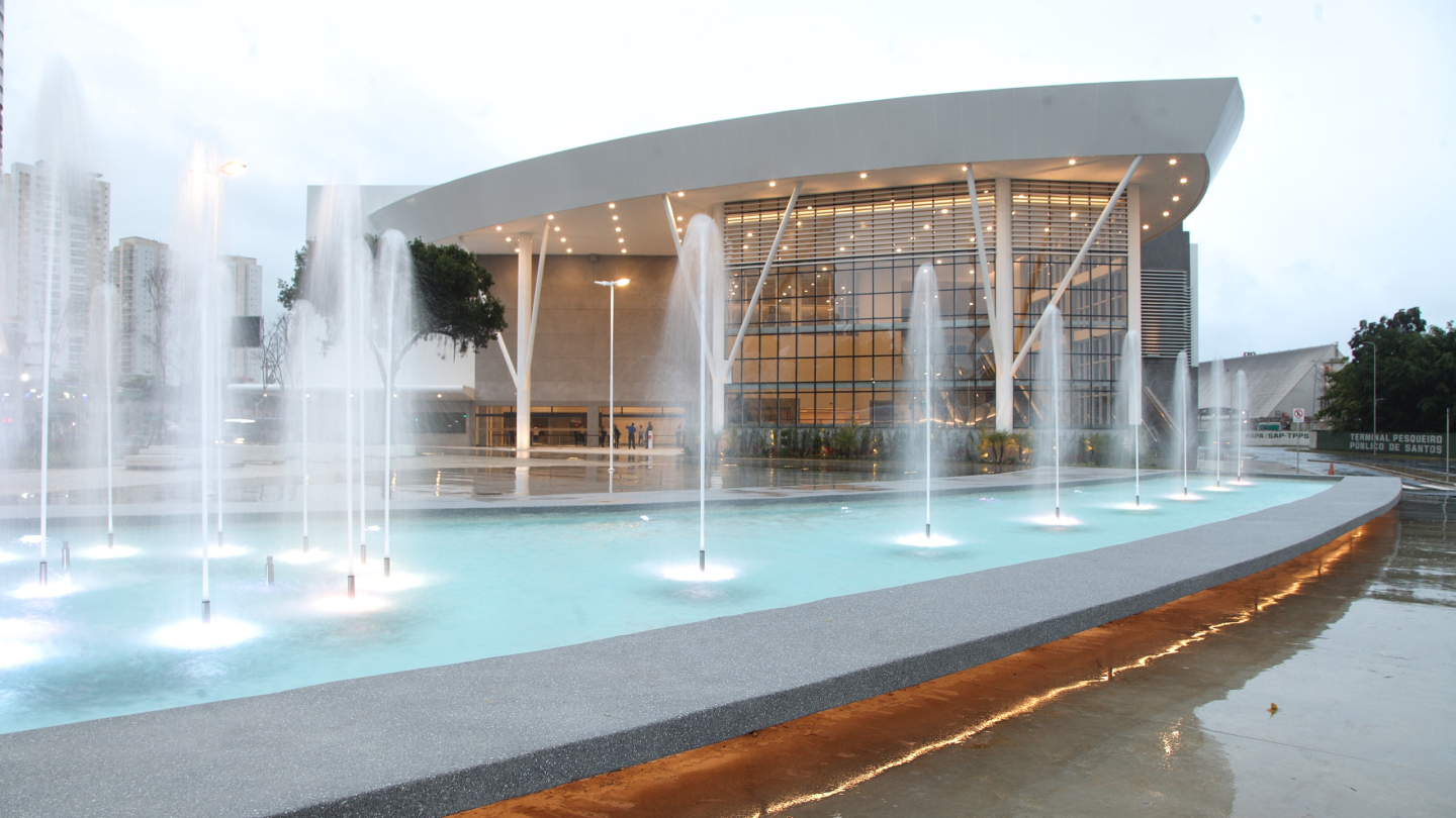 A imagem mostra a fachada do Santos Convention Center, localizado em Santos. O prédio tem uma estrutura moderna e imponente, com grandes janelas de vidro que permitem a entrada de luz natural, criando uma sensação de amplitude e transparência. O design é contemporâneo, com linhas retas e uma fachada clean, destacando-se pela combinação de materiais como vidro e concreto.