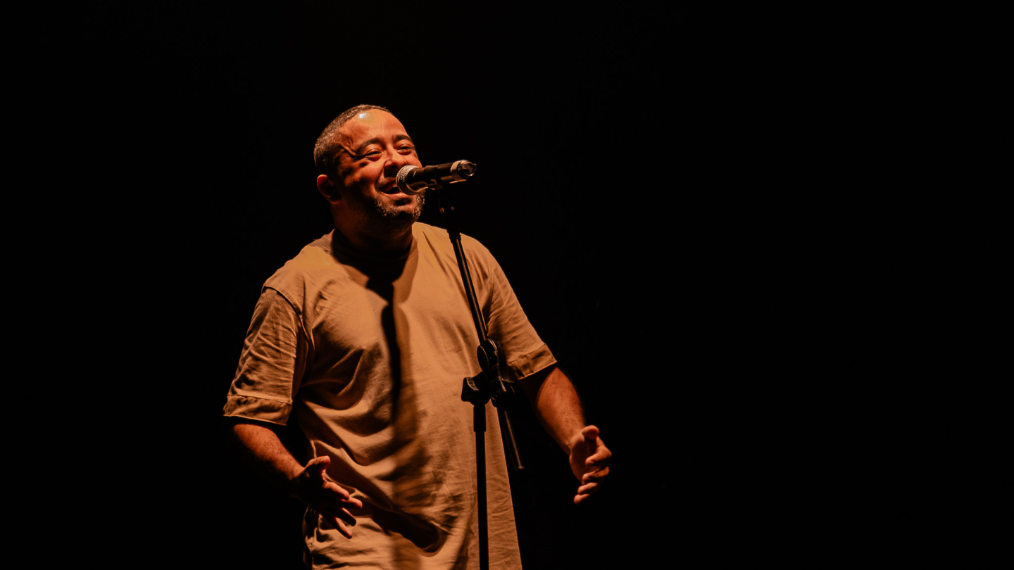 Um homem canta no palco com microfone, iluminado por uma luz quente em meio ao fundo escuro